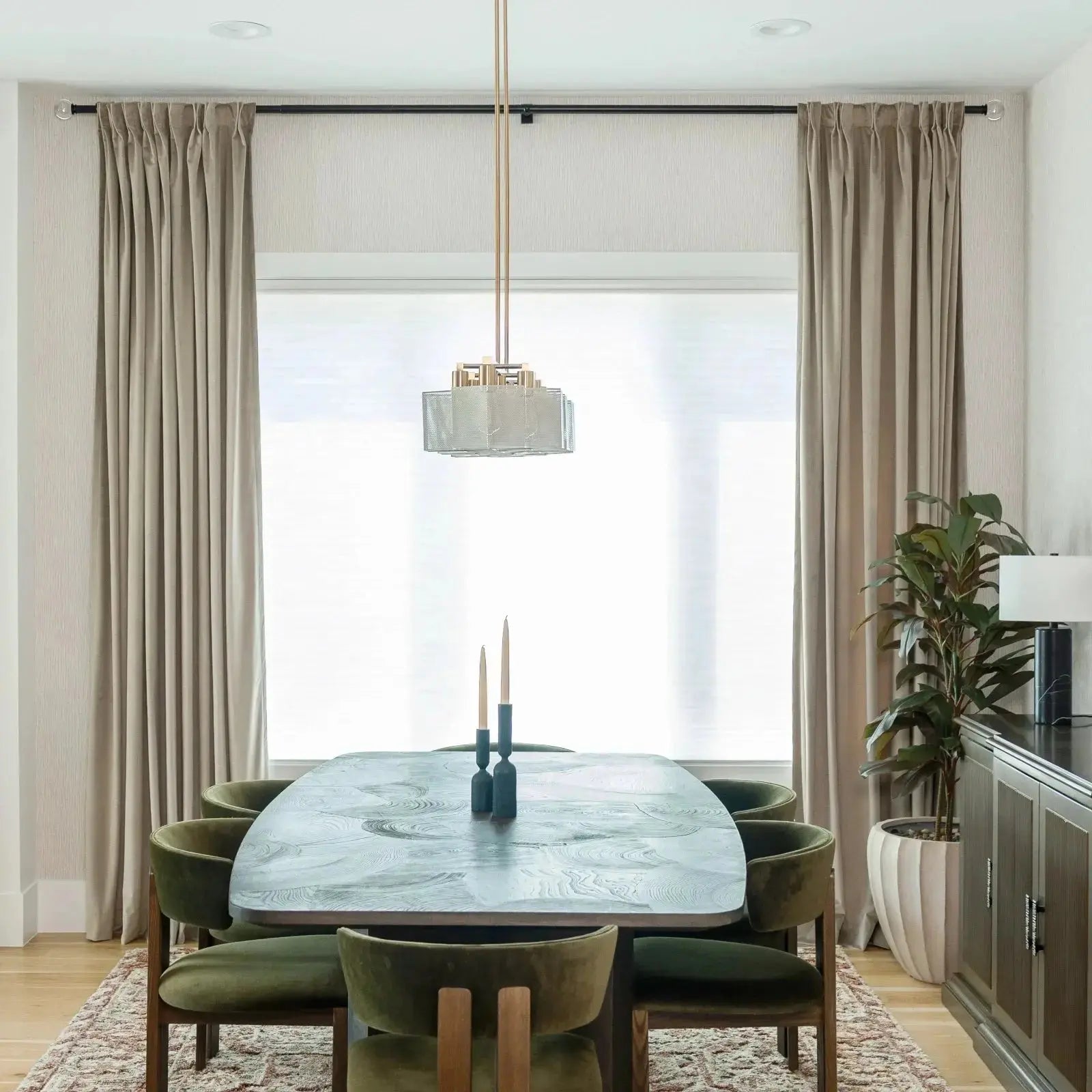 A modern dining room interior showcases an oval grey wood grain patterned dining table surrounded by moss-green velvet chairs, illuminated by a geometric gold and mesh pendant light, with dark teal candlestick holders on the table and a large ficus plant in a ribbed white planter to the right