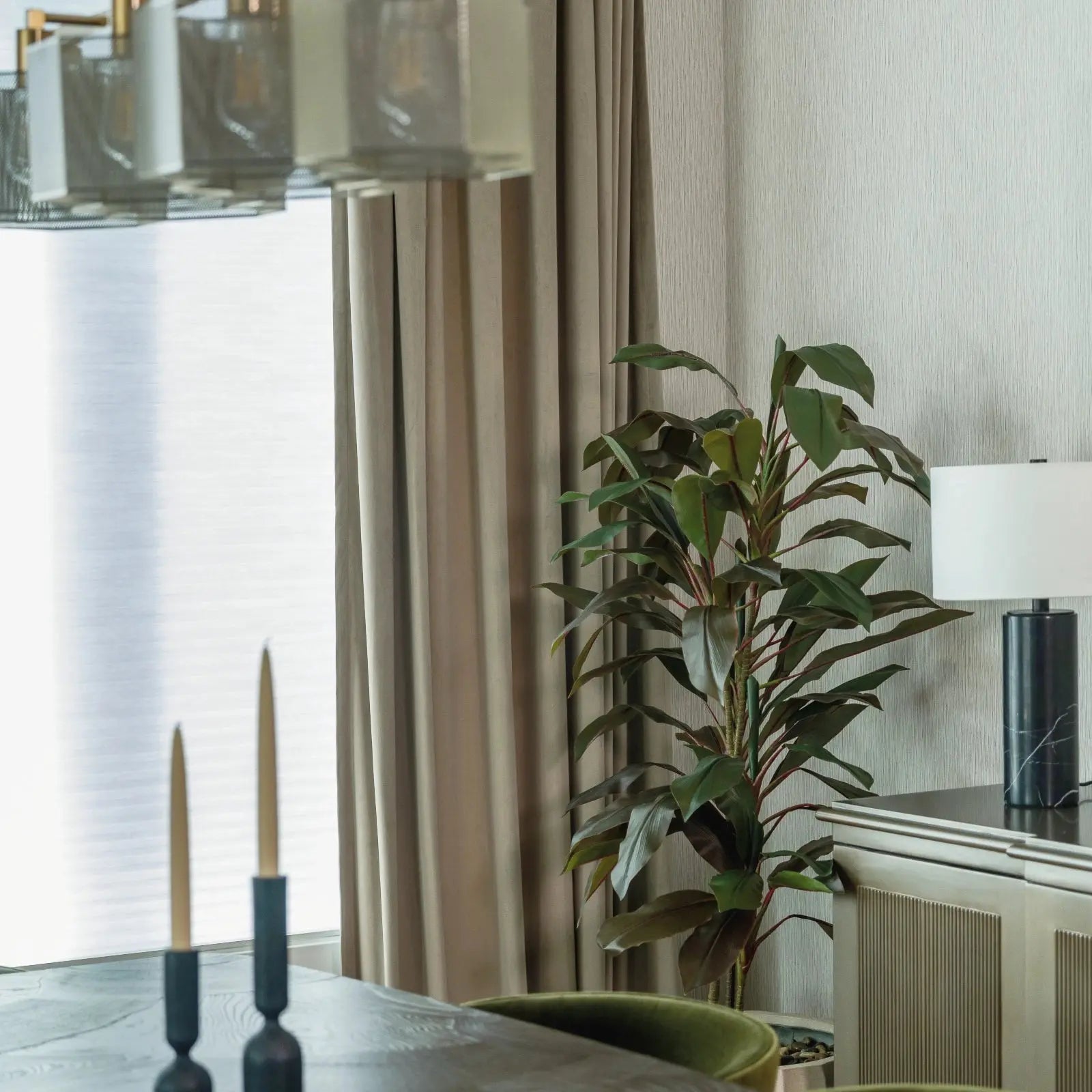 A modern dining room interior showcasing a geometric linear chandelier, gold tapered candles on a dark table, and a black marble table lamp with a tall green plant