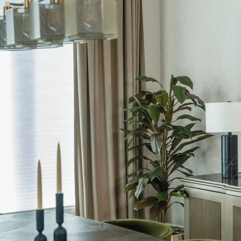 A modern dining room interior showcasing a geometric linear chandelier, gold tapered candles on a dark table, and a black marble table lamp with a tall green plant