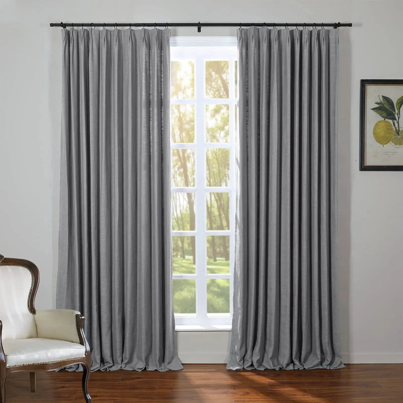 Photograph of a modern room with elegant grey pinch-pleat curtains on a black rod, framing a multi-pane window looking out to sunlit green trees, beside a vintage cream upholstered armchair and a framed lemon botanical print on the wall