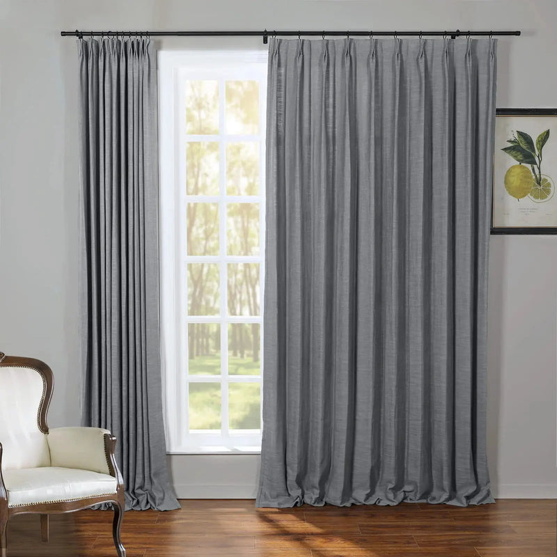 A well-lit interior room features a tall white-paned window framed by textured grey pinch-pleat drapes a classic white armchair with wood accents and a framed botanical print of yellow lemons all set on a warm hardwood floor looking out to bright green trees