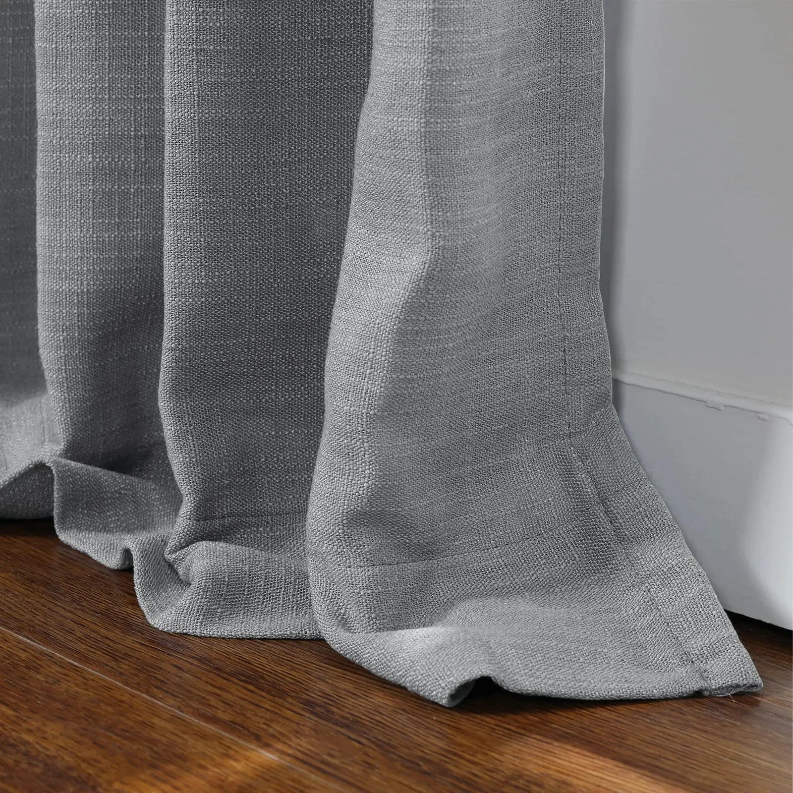 A close-up of grey linen-look pleated curtains pooling on a dark brown hardwood floor against a white baseboard