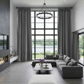 A modern grey living room with a floor-to-ceiling grid window offering a serene lake view includes a large grey sectional sofa with checkered pillows a circular black chandelier and a vase of vibrant purple flowers on the coffee table
