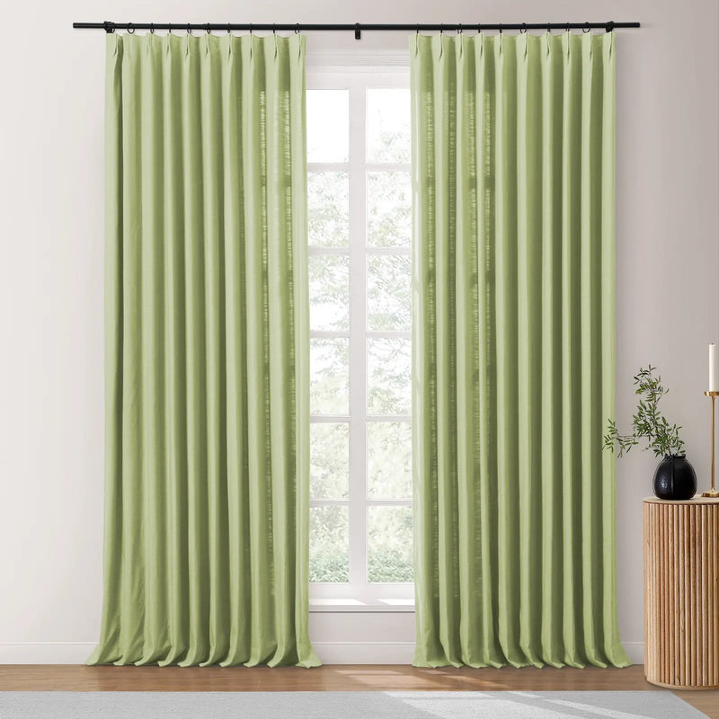 Digital image of light green pinch pleat curtains with a linen texture hung on a black rod against a bright window, next to a fluted wooden table with a black vase and foliage