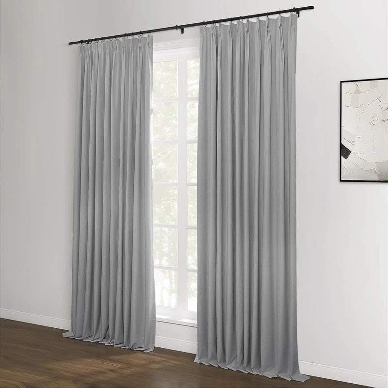 A contemporary room interior featuring long grey pleated curtains on a black rod, framing a multi-paned window, with dark wood flooring and a minimalist black and white abstract artwork on the adjacent white wall