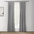 A pair of gray rod pocket curtains hang on a black curtain rod in a brightly lit room featuring a multi-paned window, an abstract white and black painting, and a wooden floor with a textured rug