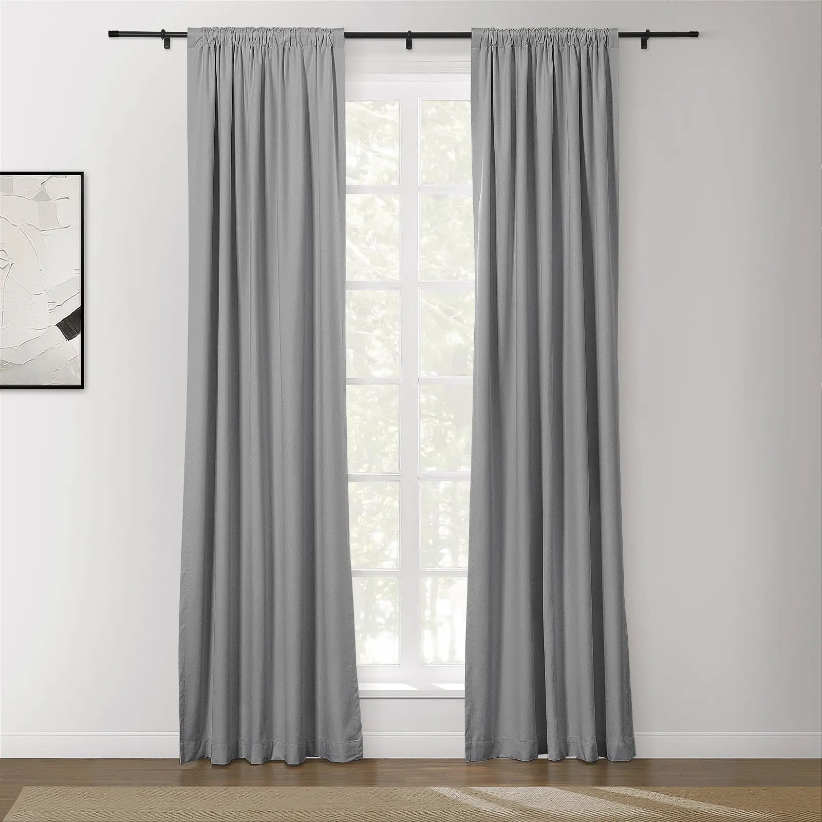 A pair of gray rod pocket curtains hang on a black curtain rod in a brightly lit room featuring a multi-paned window, an abstract white and black painting, and a wooden floor with a textured rug