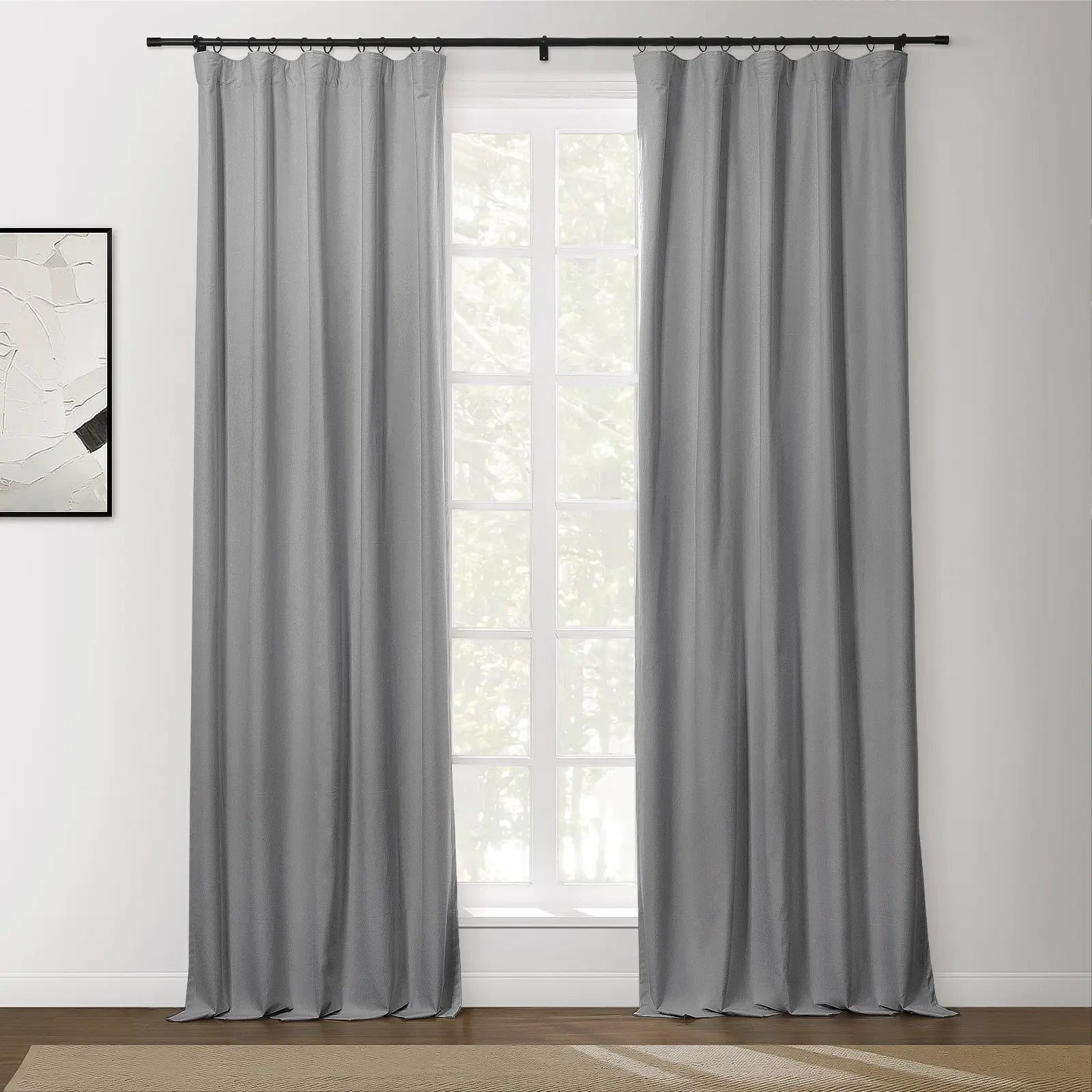 A pair of gray textured curtains hangs from a black rod in an interior room, framing a multi-pane window with green foliage outside, and abstract white and black artwork on the left wall