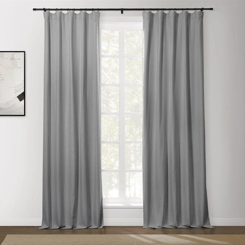 A pair of gray textured curtains hangs from a black rod in an interior room, framing a multi-pane window with green foliage outside, and abstract white and black artwork on the left wall
