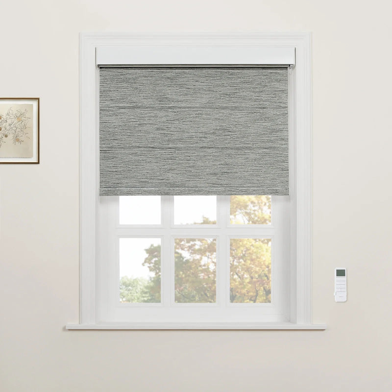 A grey textured roller Blind installed on a white multi-pane window, with a wall-mounted remote control and a small framed botanical drawing on the light wall