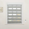A window in an interior room featuring white trim, zebra-style Blinds with alternating grey textured and sheer white stripes, a wall-mounted white remote control, and a framed botanical print on the left