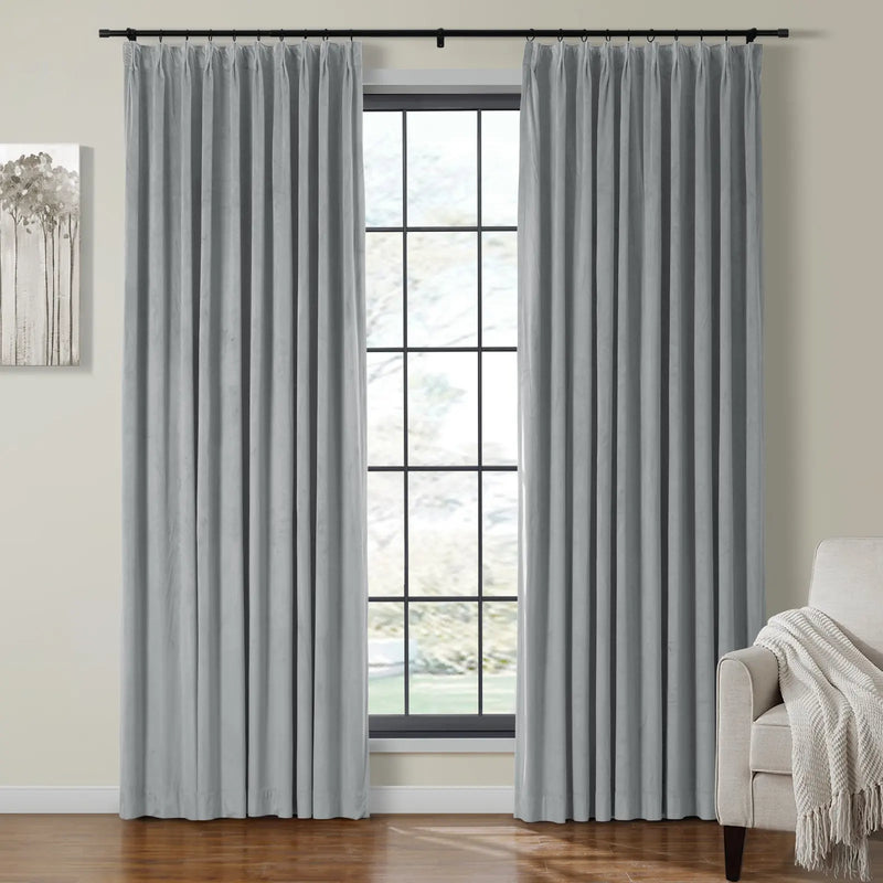A modern interior room showcasing elegant grey pleated velvet curtains on a black rod flanking a grid-paned window, complemented by a beige armchair with a white textured throw blanket and abstract tree wall art