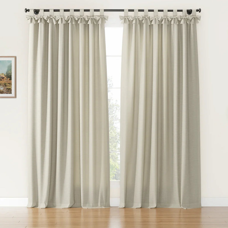 A pair of natural textured beige tab-top curtains featuring decorative bows and a black rod in a sunlit room with a small framed landscape painting