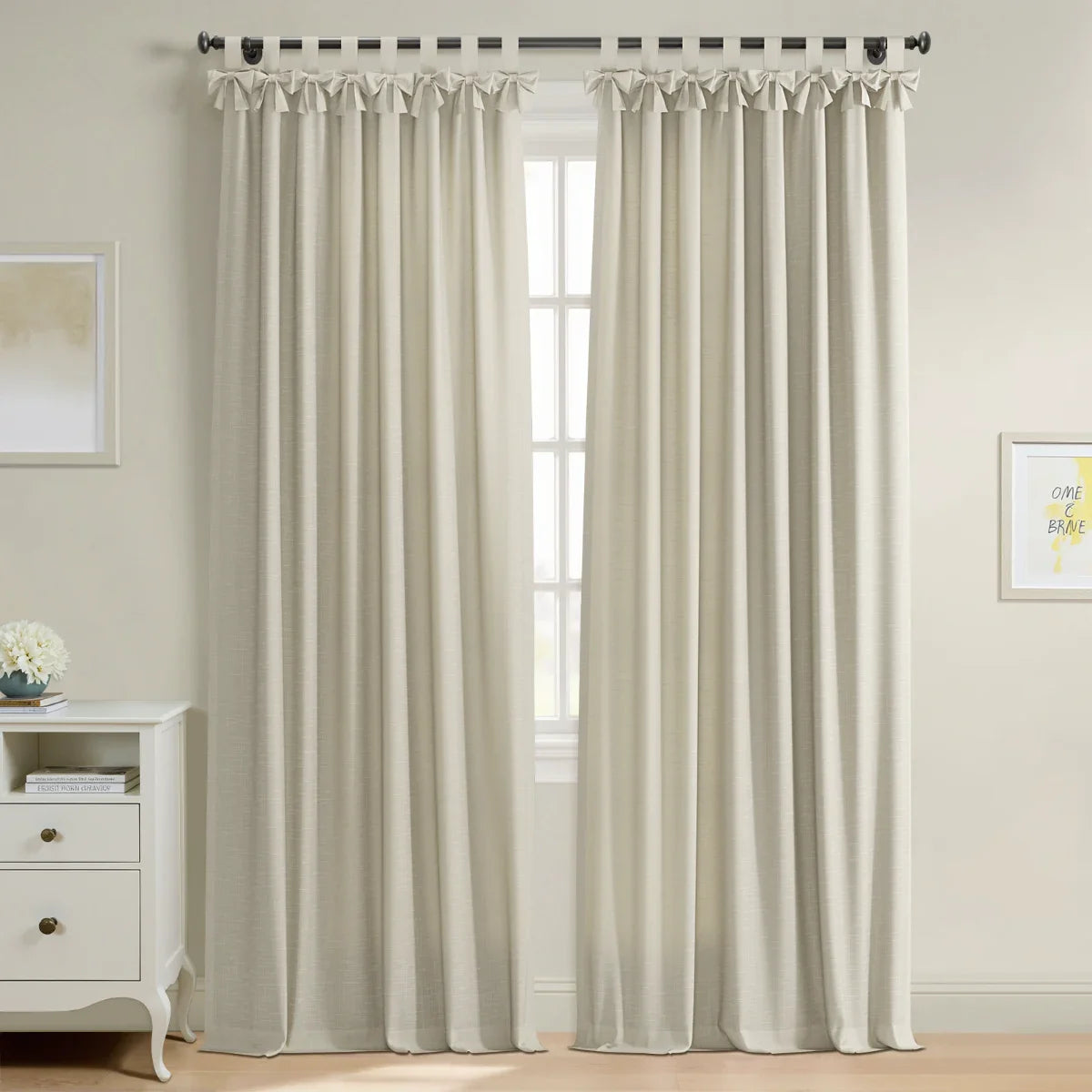Light beige linen-look curtains with decorative tie-top tabs cover a window in a bright room featuring a white dresser with a blue vase of flowers and framed yellow inspirational artwork