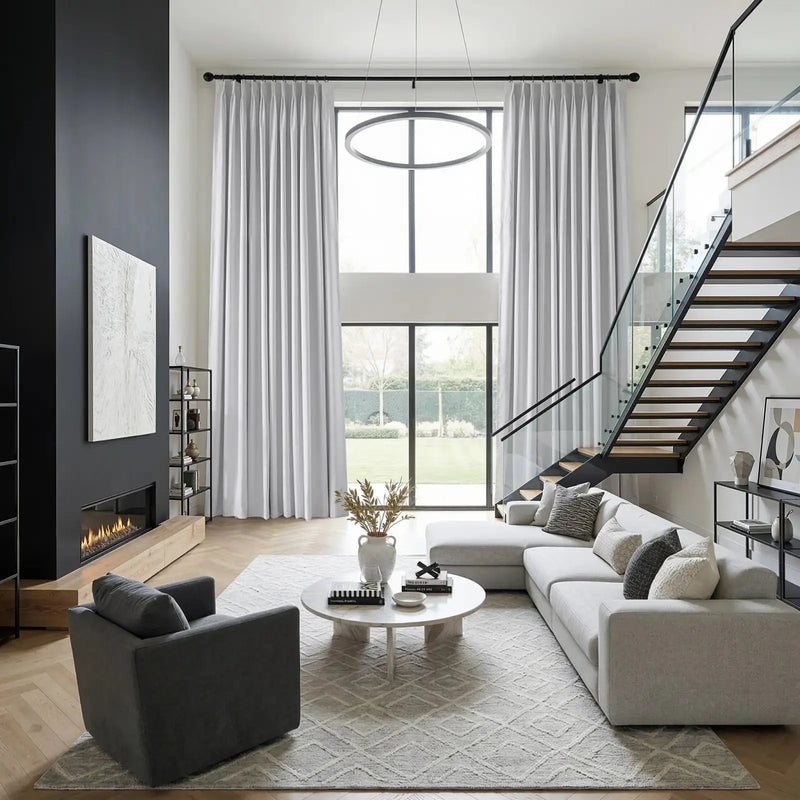 Sleek modern living room interior with a black fireplace wall featuring white abstract art, towering windows with light gray drapes, a minimalist circular chandelier, and a dramatic floating wooden and glass staircase