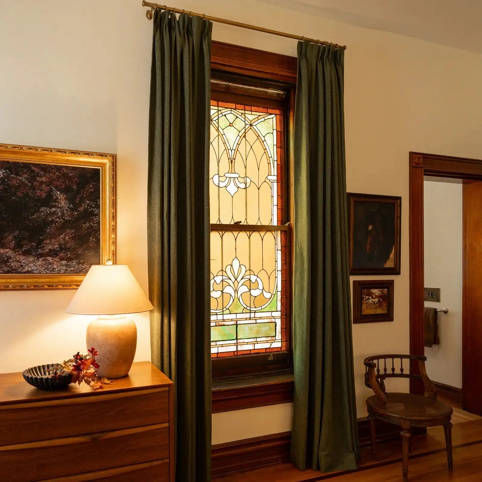 Photograph of an elegant room interior with an ornate arched stained glass window flanked by olive green curtains, a wooden dresser adorned with a glowing ceramic lamp and fall foliage, and framed art on the wall