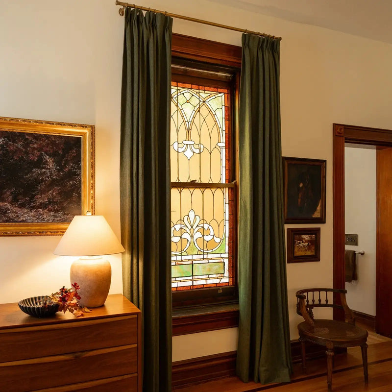 Photograph of an elegant room interior with an ornate arched stained glass window flanked by olive green curtains, a wooden dresser adorned with a glowing ceramic lamp and fall foliage, and framed art on the wall