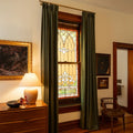 Photograph of an elegant room interior with an ornate arched stained glass window flanked by olive green curtains, a wooden dresser adorned with a glowing ceramic lamp and fall foliage, and framed art on the wall