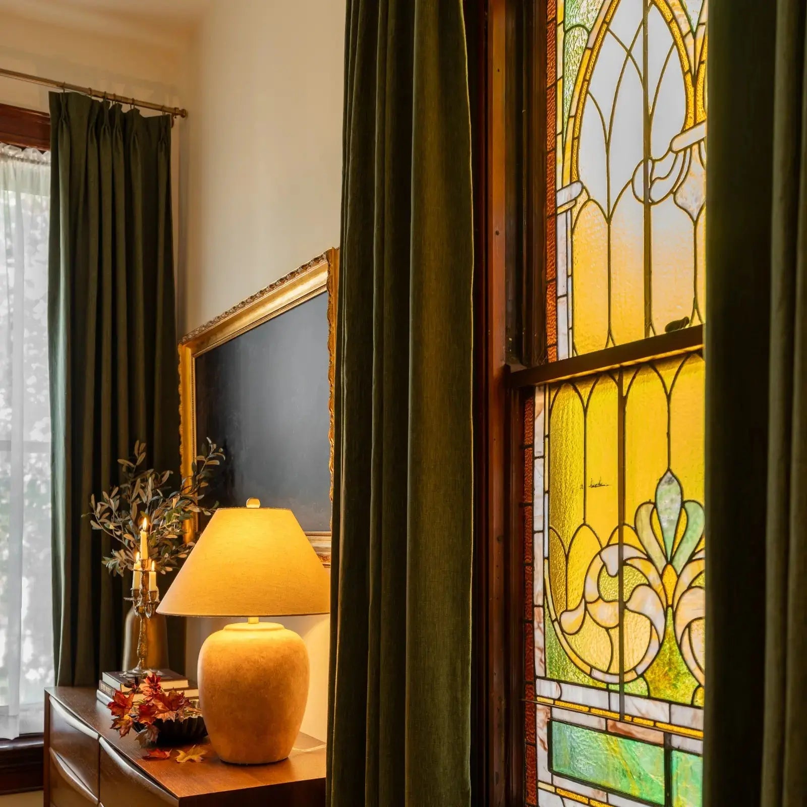 An elegant interior room featuring olive green velvet curtains a vibrant yellow and green art nouveau stained glass window a warm glowing ceramic lamp on a dark wooden dresser adorned with autumnal leaves and a gold-framed dark mirror