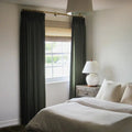 A photograph of a modern bedroom interior showcasing dark green pleated drapes on a gold rod, a natural woven wood blind, and a white ceramic globe lamp on a rustic nightstand