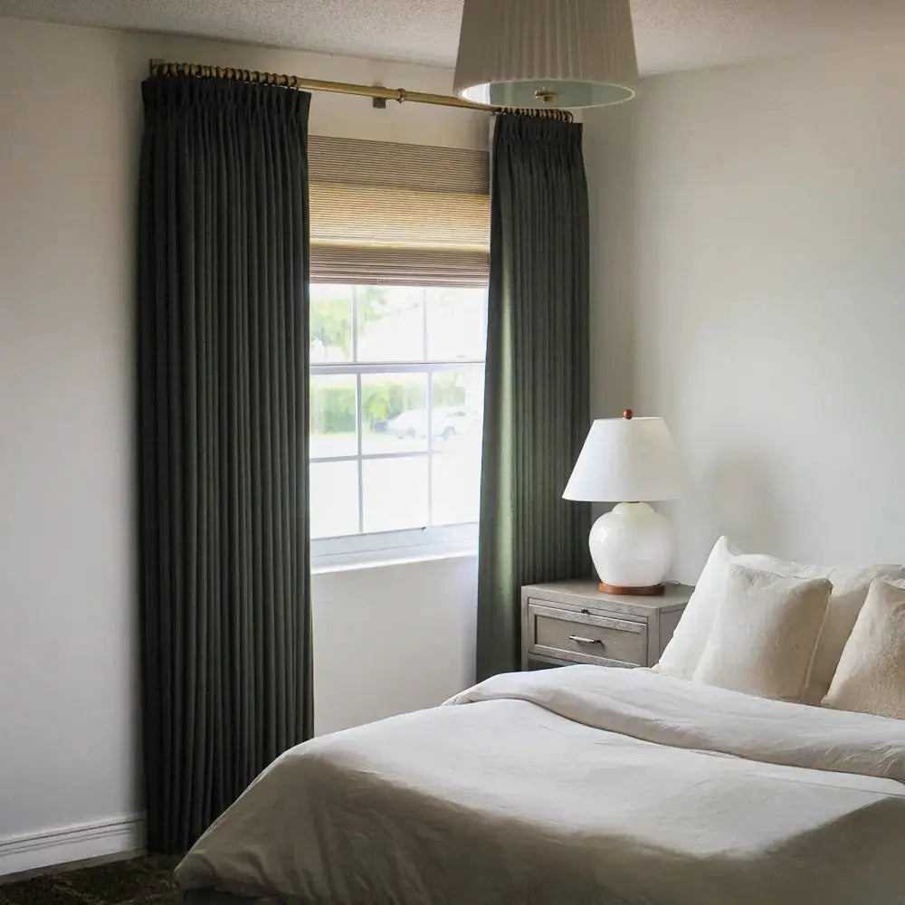 A photograph of a modern bedroom interior showcasing dark green pleated drapes on a gold rod, a natural woven wood blind, and a white ceramic globe lamp on a rustic nightstand