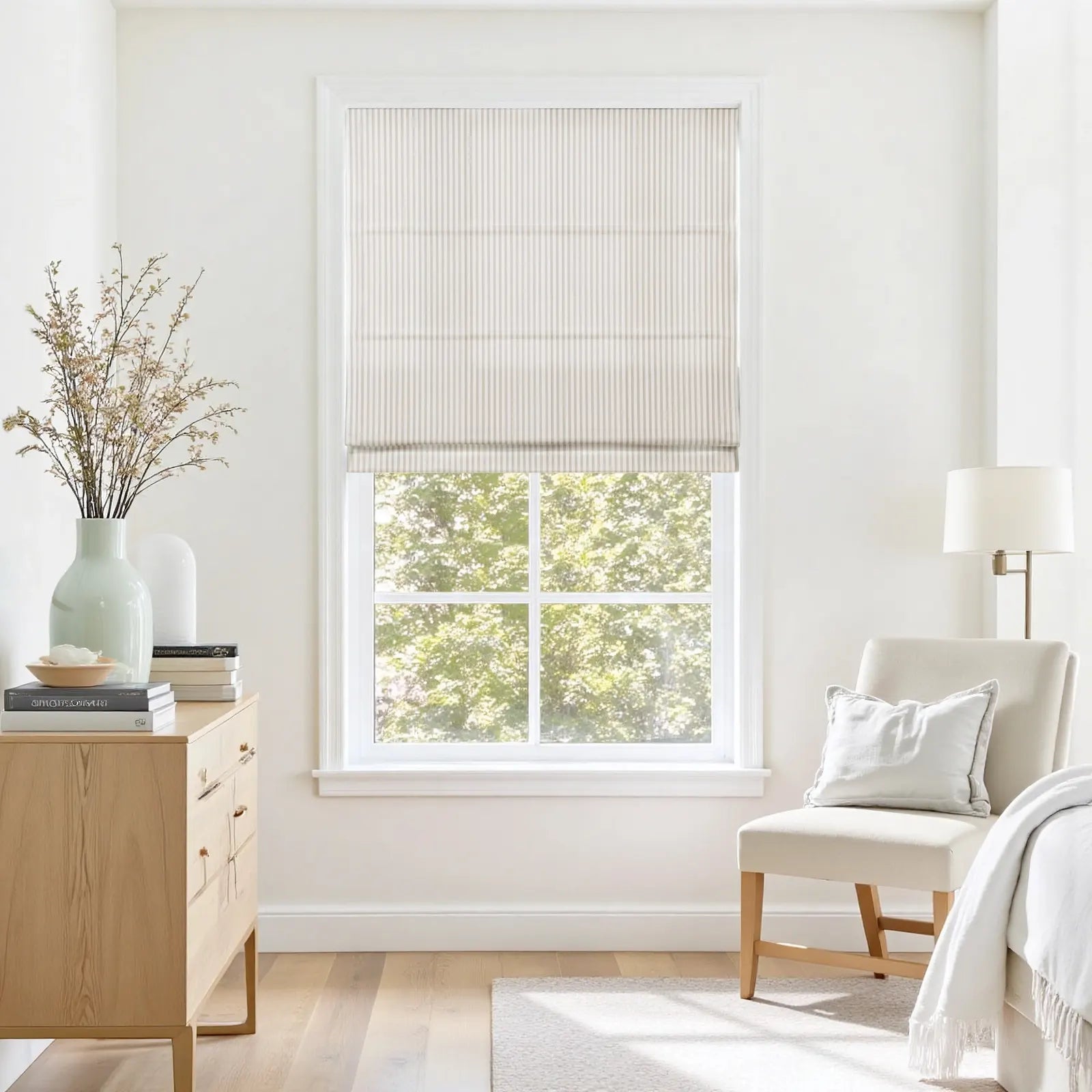 Photography of a serene bedroom interior featuring a beige striped Roman shade, a window overlooking green trees, a light wood dresser with a mint green vase of dried botanicals, and a cream upholstered chair