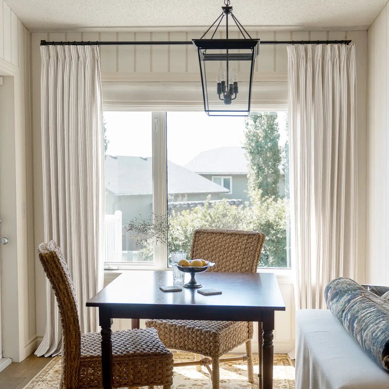 Photograph of a cozy dining area in a bright room featuring a dark wood table with woven wicker chairs, a black lantern chandelier, and striped curtains flanking a large window revealing a sunny suburban view, with a bowl of lemons and dried branches on the table