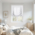 A bright modern minimalist bedroom featuring white hobbled Roman shades, light wood furniture, a bed with white linens and a grey throw, and dried yellow floral arrangements
