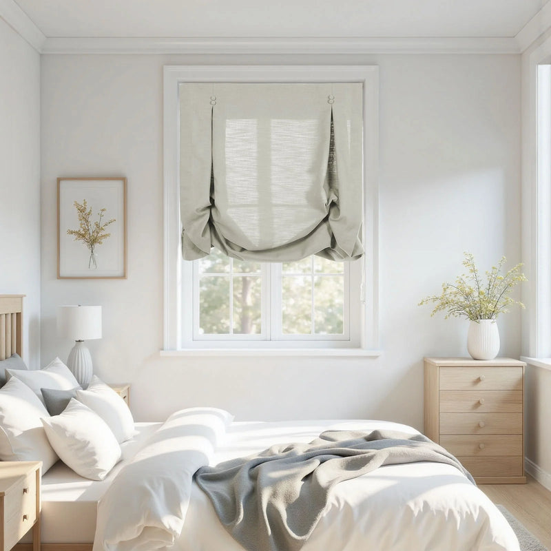 A bright minimalist bedroom interior featuring a bed with crisp white linens and a grey throw, light wood furniture, a window with a stylish light grey linen Roman shade, and delicate yellow wildflowers in a framed print and a vase
