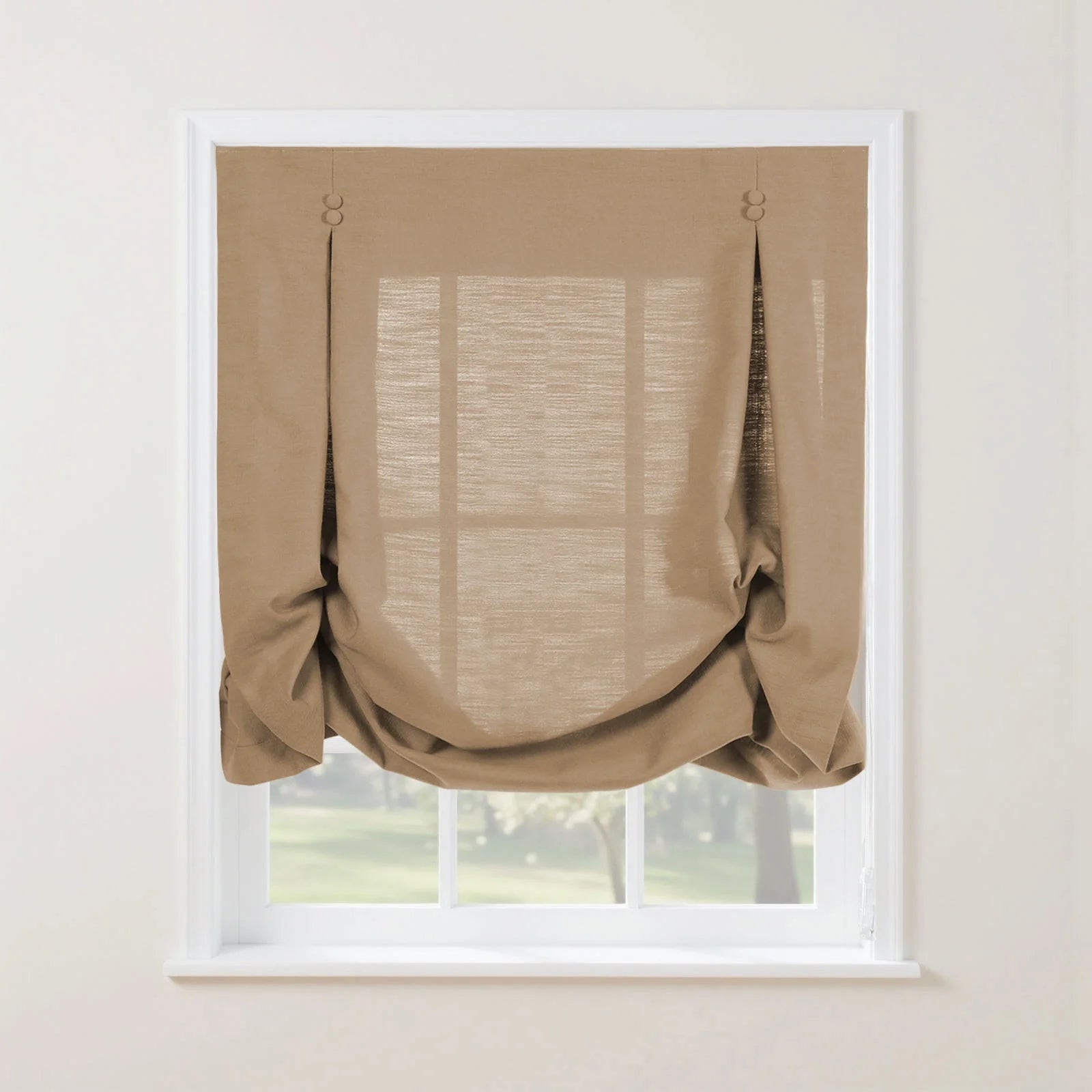 A medium brown linen-look relaxed Roman shade with two decorative button accents and soft bottom folds partially covers a white-framed window overlooking a green outdoor landscape