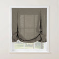 A medium grey linen blend Roman shade featuring soft bottom gathers and two decorative button-loop details on a white window frame, showing a faint view of a green outdoor garden
