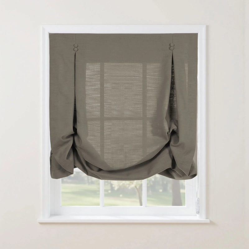 A medium grey linen blend Roman shade featuring soft bottom gathers and two decorative button-loop details on a white window frame, showing a faint view of a green outdoor garden