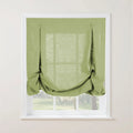 An apple green linen tie-up window shade with two decorative fabric loops and a softly gathered bottom adorns a clean white interior window frame, offering a filtered view of a bright outdoor green landscape
