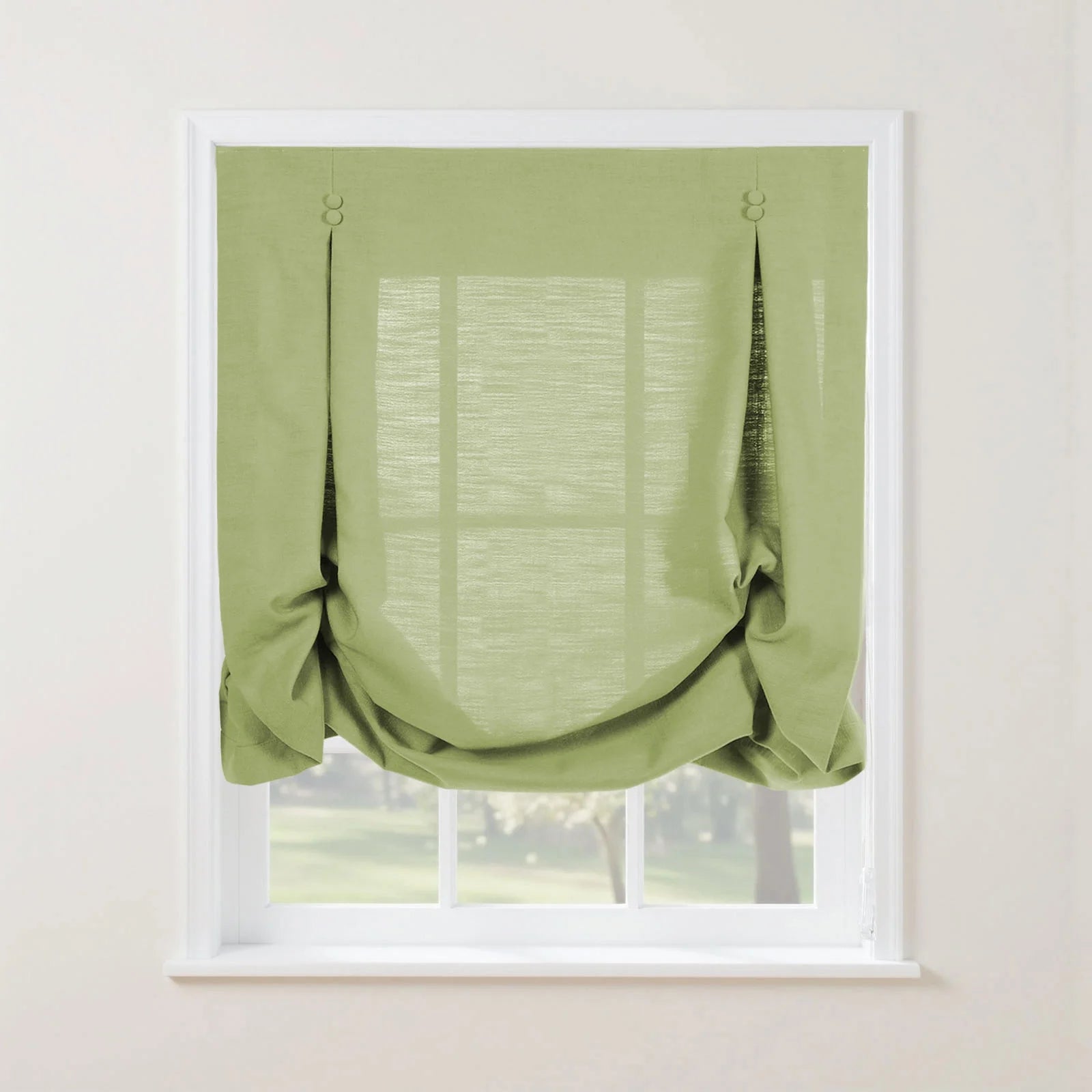 An apple green linen tie-up window shade with two decorative fabric loops and a softly gathered bottom adorns a clean white interior window frame, offering a filtered view of a bright outdoor green landscape