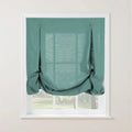 A teal linen balloon shade on a white window frame, featuring decorative buttoned pleats at the top corners and soft gathers, partially revealing a blurred green outdoor scene