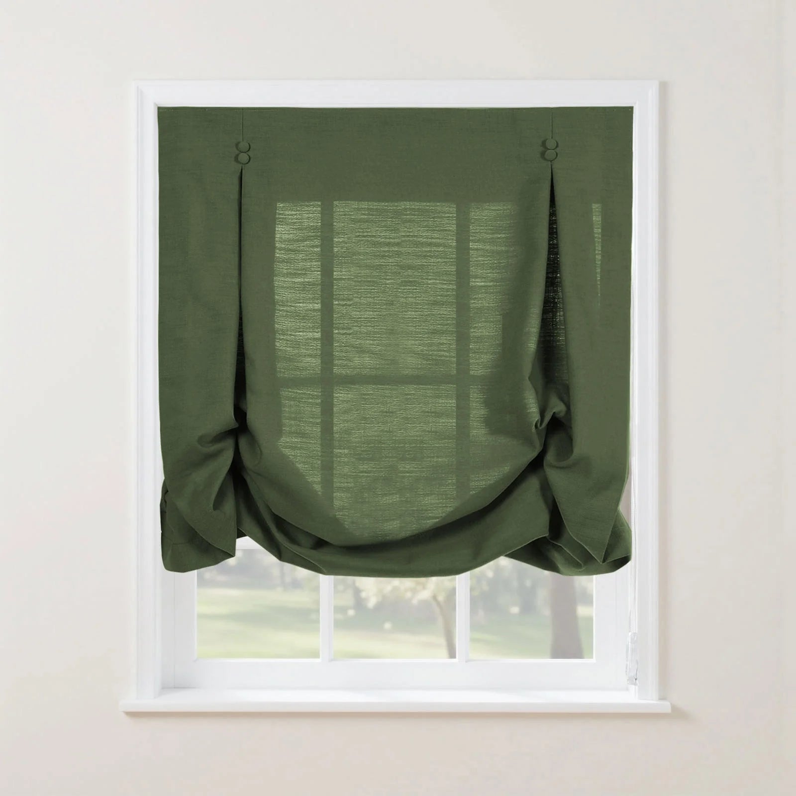 A forest green linen tie-up Roman shade with decorative button tabs and soft bottom folds partially obscures a white window frame overlooking a sunlit garden