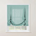 Light teal linen tie-up Roman shade featuring decorative buttons at the top and a gathered bottom revealing a verdant outdoor landscape through the window