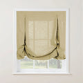 A light beige linen tie-up Roman shade with decorative loops covers a white-framed window revealing soft light and green outdoor scenery