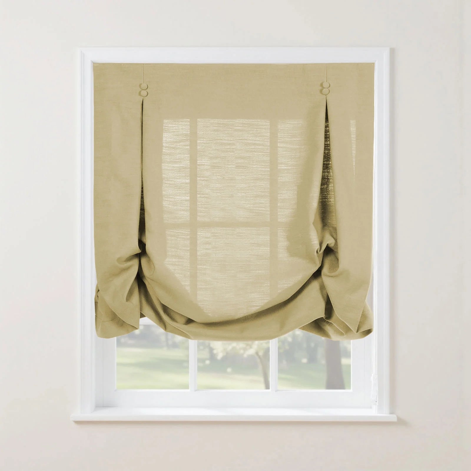 A light beige linen tie-up Roman shade with decorative loops covers a white-framed window revealing soft light and green outdoor scenery