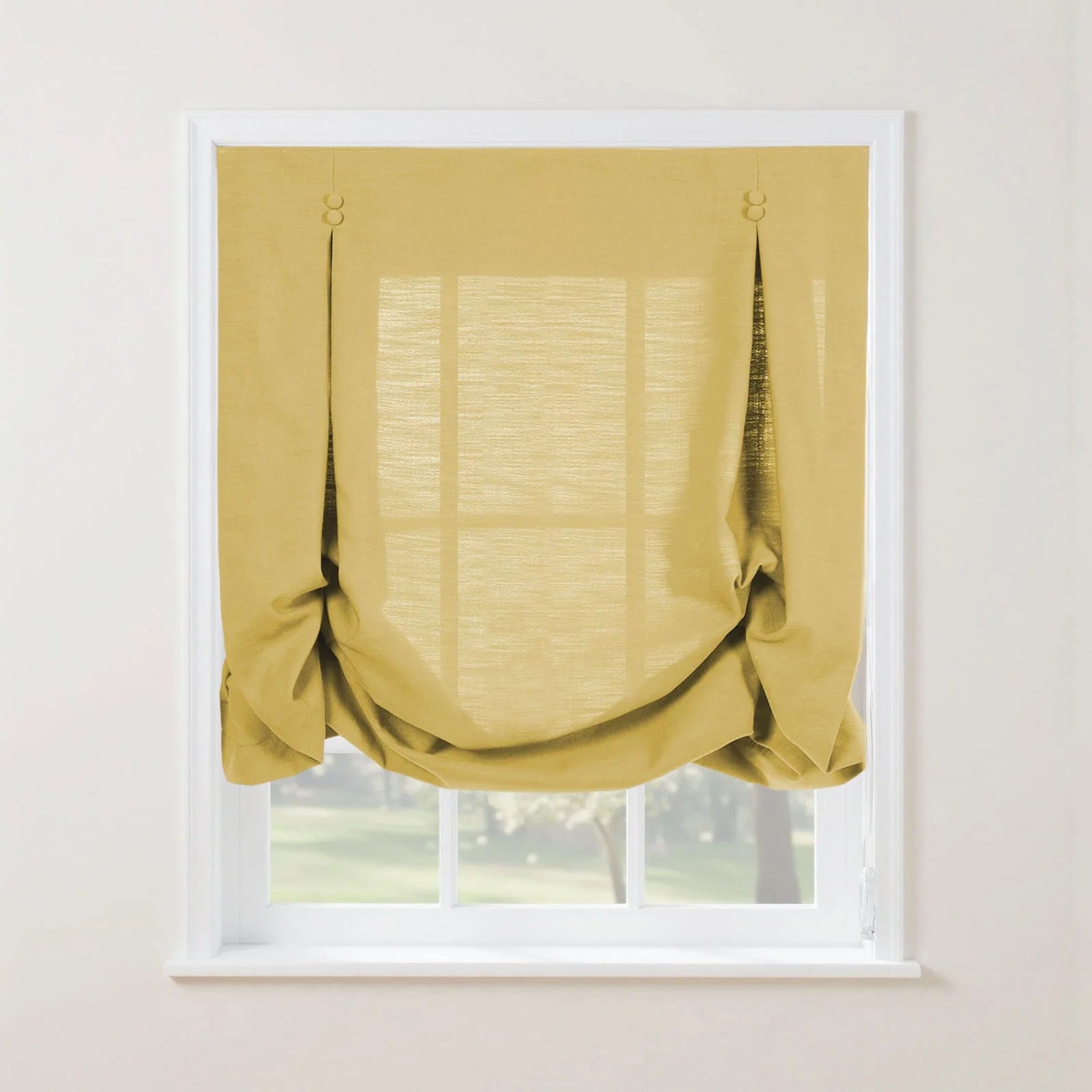 A golden yellow linen-textured tie-up Roman shade featuring decorative loops partially covers a white-framed window overlooking blurred green trees