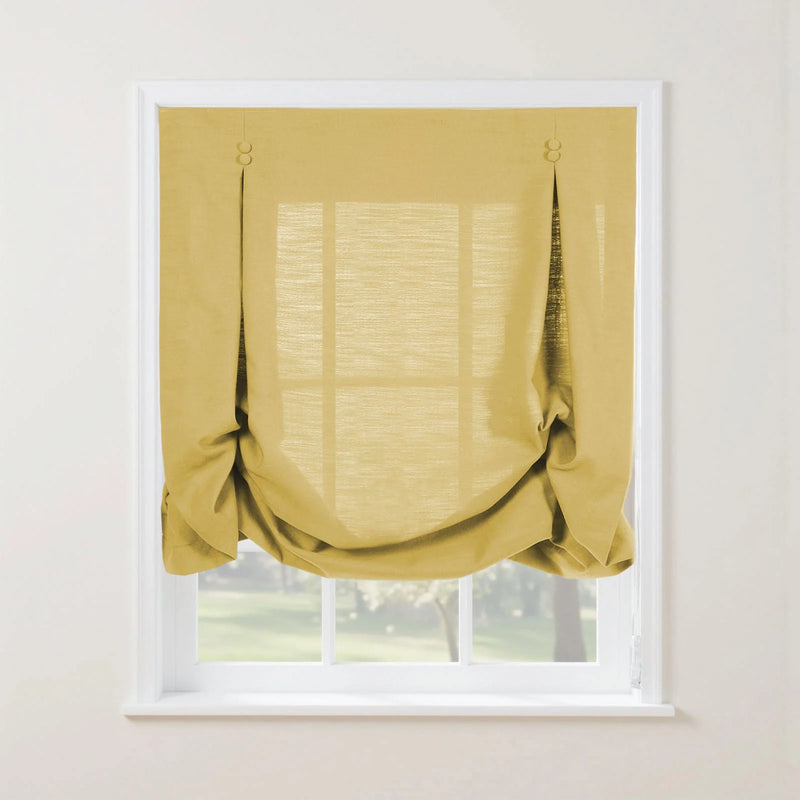 A golden yellow linen-textured tie-up Roman shade featuring decorative loops partially covers a white-framed window overlooking blurred green trees