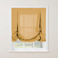 A mustard yellow textured relaxed Roman shade with decorative button loops pulled up creating soft folds against a white window frame revealing a blurry green outdoor landscape