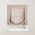 Photograph of a light beige textured linen relaxed roman shade with decorative button details and soft bottom folds, covering a white paned window with a blurred green garden view