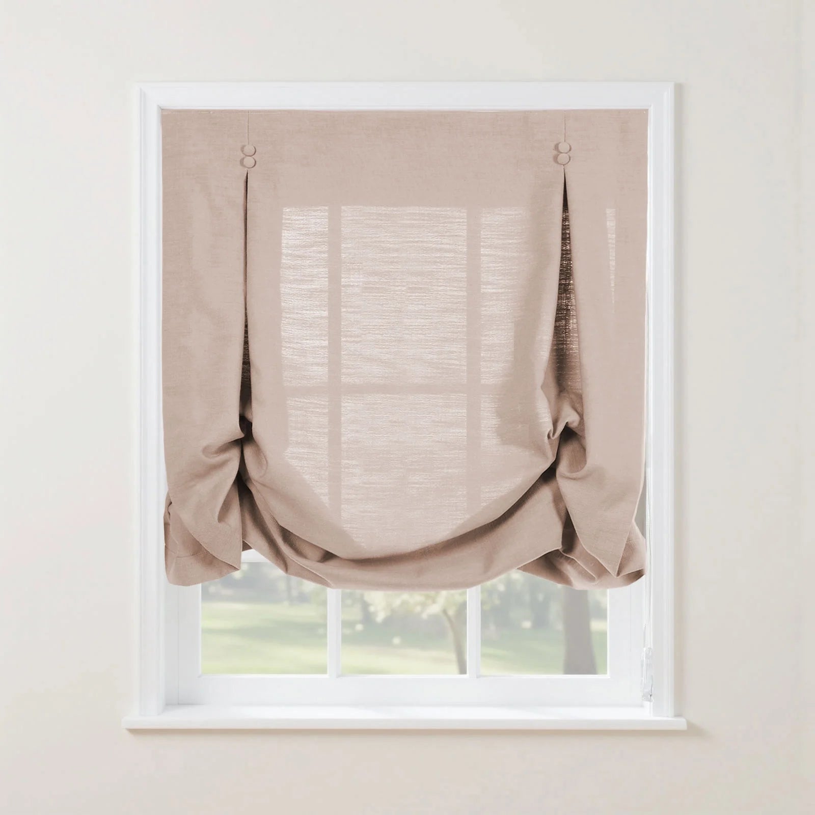 Photograph of a light beige textured linen relaxed roman shade with decorative button details and soft bottom folds, covering a white paned window with a blurred green garden view