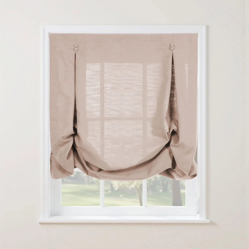 Photograph of a light beige textured linen relaxed roman shade with decorative button details and soft bottom folds, covering a white paned window with a blurred green garden view