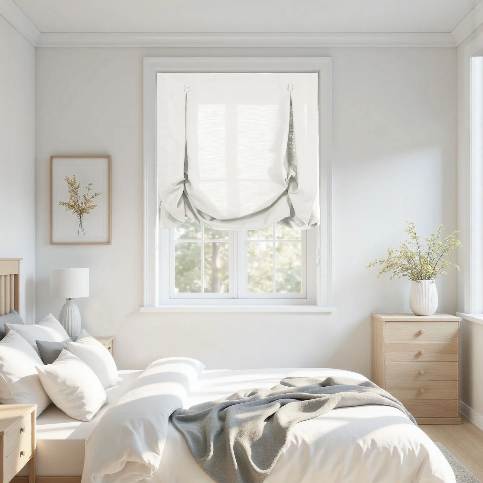 Photograph of a bright minimalist bedroom interior featuring white Roman shades over a window, a bed with white bedding and a gray throw, light wood nightstands, a framed yellow floral print, and a white vase with dried yellow flowers