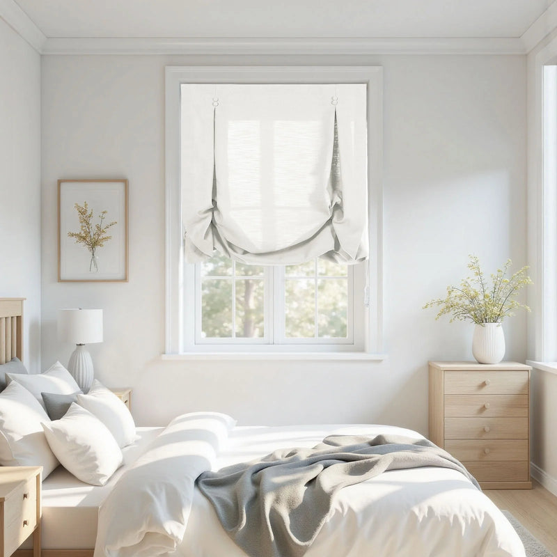 Photograph of a bright minimalist bedroom interior featuring white Roman shades over a window, a bed with white bedding and a gray throw, light wood nightstands, a framed yellow floral print, and a white vase with dried yellow flowers