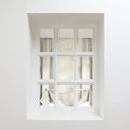 Photography of a minimalist white nine-pane window set in a deep recessed wall, featuring light beige roman blinds partially tied with white cords