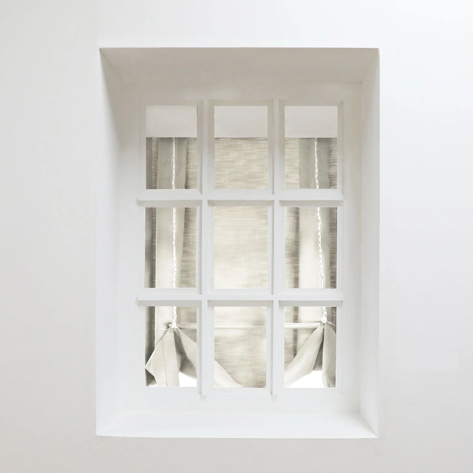 Photography of a minimalist white nine-pane window set in a deep recessed wall, featuring light beige roman blinds partially tied with white cords