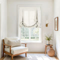 A bright interior room featuring a natural wood armchair with cream cushions, a window with a white linen balloon shade, a wooden side table displaying green and yellow foliage in a ceramic vase, a woven rattan basket, a brass wall sconce, and framed abstract art